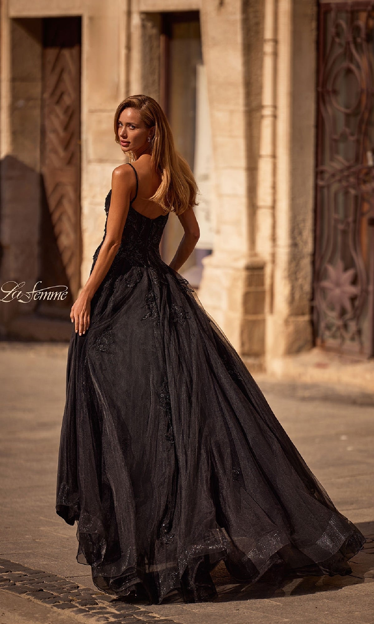 Wearing the La Femme 32781 Long Black Glitter Prom Dress, a woman stands outdoors, gazing over her shoulder with historic stone buildings as the scenic backdrop.
